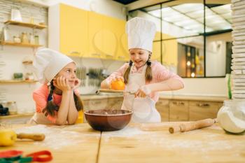 Two little girls cooks in caps rubs orange to the bowl, cookies preparation on the kitchen. Kids cooking pastry, children chefs preparing cake