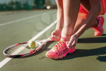 Female tennis player tying laces on sneakers, racket and ball on the ground. Summer season active sport game on open air