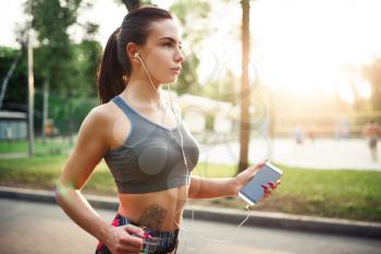 Athletic girl jogging with headphones on walkway in summer park. Woman on outdoors morning workout