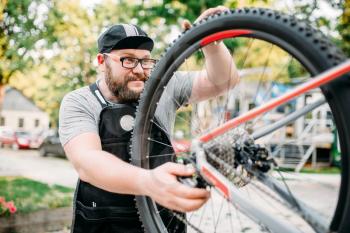 Bicycle repairman works with bike wheel, cycle workshop outdoor. Bearded mechanic in apron