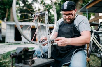 Male person in apron adjusts bike spokes and wheel with service tools. Cycle workshop outdoor. Bicycling sport, professional bicycle mechanic