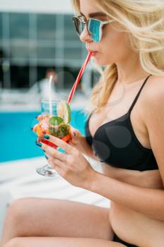 Slim woman in swimsuit sitting by the poolside and drinks cocktail. Happy girl relax near the swimming pool. Summer vacations
