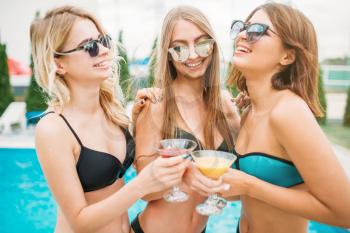 Three happy girls in swimsuits and sunglasses drinks cocktails and have fun near the swimming pool. Resort holidays. Tanned women on summer holidays