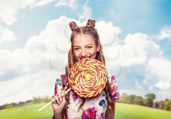 Beautiful young woman with playful look bites huge candy. Stylish girl with blonde curly hair. Stylish girl in colorful summer dress, sunny meadow on background.