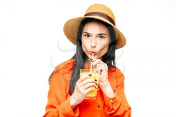 Cute woman in shirt and straw hat drinks freshly squeezed orange juice, white background. Young girl with yellow beverage, healthy lifestyle