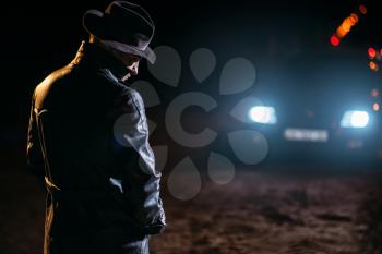 Serial maniac in black leather coat and hat, back view, car light at the night on background. Horror, bloody butcher