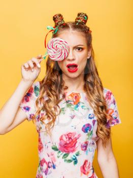 Beautiful young woman holds in hand candy, emotion of indignation on her face. Stylish girl in summer colorful dress. Studio portrait, yellow wall on background.