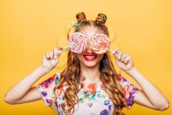 Amazing cute young girl on the yellow background holding huge sweets. Stylish girl in summer colorful dress, studio portrait. Portrait of attractive lady with two big lollypop.