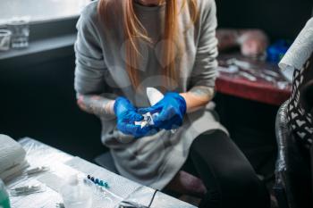 Female tattooist in blue sterile gloves prepares tattoo machine, master in salon. Professional tattooing in studio