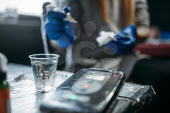 Female tattooist hands in blue sterile gloves holds tattoo machine, professional work tools on background. Tattooing in salon