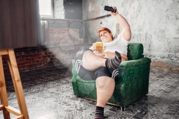 Overweight woman sitting in chair and drinks beer, laziness and obesity. Unhealthy lifestyle, fatty female