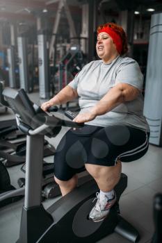 Fat woman training on exercise bike in gym. Calories burning, obese female person in sport club