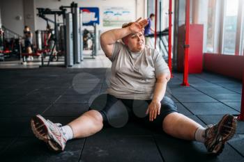 Tired overweight woman sits on the floor in gym. Calories burning, obese female person in sport club, fat people