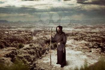 Medieval monk in black robe with hood is walking in the desert