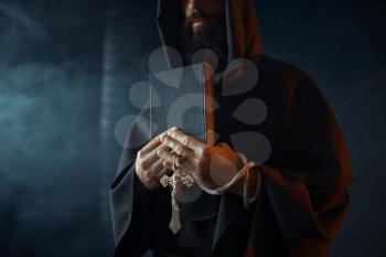 Medieval monk holds book and wooden cross in hands, black background, secret ritual. Mysterious friar in dark cape. Mystery and spirituality