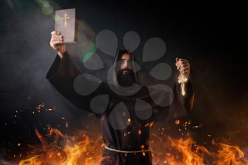 Medieval monk stands in fire with book in hands in fire, black background, secret ritual. Mysterious friar in dark cape. Mystery and spirituality