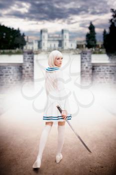 Anime style blonde girl with sword against ancient castle, back view. Cosplay woman, asian culture, doll with blade in cold tones, night cityscape on background
