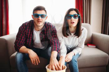 Bewitched couple in glasses sitting on couch, man and woman watch tv and eats popcorn at home, window on background