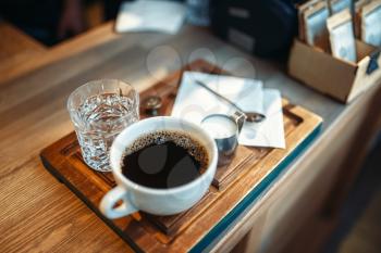 Cup of fresh black coffee, milk and glass of water stands on wooden counter in espresso cafe, cafeteria interior on background, nobody