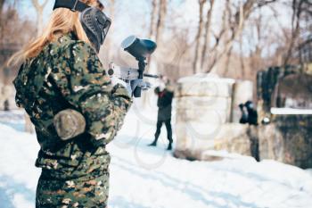 Female paintball player shooting at the enemy, back view, winter forest battle. Extreme sport game, players in protection masks and camouflage lies on the ground and holds weapons in hands
