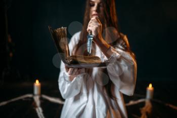 Witch in white shirt holds knife and reads spell, pentagram circle with candles, dark magic ritual process. Occultism and exorcism