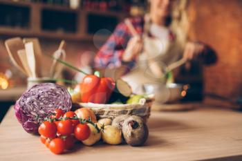 Fresh vegetables on wooden table, female cook in apron on the kitchen on background. Housewife making healthy vegetarian food, salad ingredients