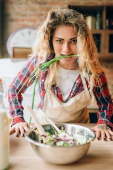 Housewife in an apron holds in teeth a brush of green onions, kitchen interior on background. Female cook making healthy vegetarian food, salad cooking