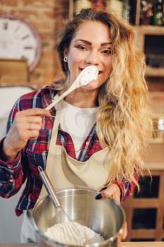 Young housewife in an apron tastes fresh dough, kitchen interior on background. Female cook prepares fresh homemade cake. Domestic pie preparation