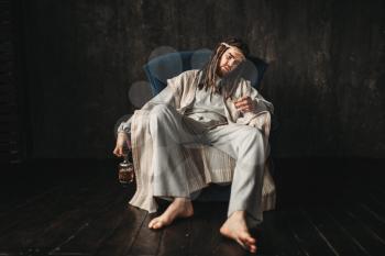 Drunken man in the image of Jesus Christ holds a stack and a bottle of alcohol sitting in a chair. Drinking is a sin concept
