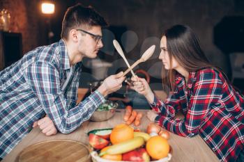 Playful young couple fight with wooden spoons on the kitchen while cooking breakfast. Man and woman preparing vegetable salad, happy family together