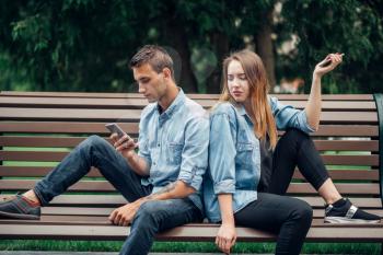 Phone addiction, young couple on the bench in park. Man and woman using their smartphones, addicted people