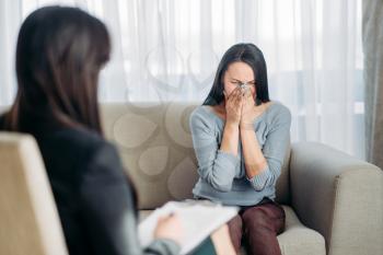 Sad female patient sitting on sofa at psychotherapist reception. Female doctor writes notes in notepad, professional psychology support