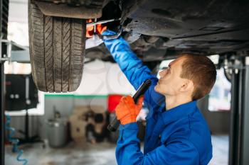 Mechanic with lamp checks the car suspension, repair station. Tire service, vehicle maintenance