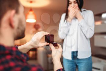 Young man makes an offer to get married to surprised woman and gives a ring. Modern apartment interior on background. Love couple relationship