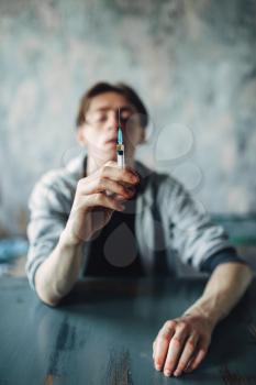 Male junkie sitting at the table with syringe in hand, grunge room interior on background. Drug addiction concept, addicted people