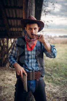 Brutal cowboy smokes a cigar, texas ranch on background, western. Vintage male person with gun on farm, wild west culture