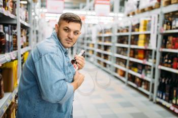 Man hides a bottle of alcohol under his shirt in supermarket. Male thief in shop, husband conducting a covert operation, shoplifling
