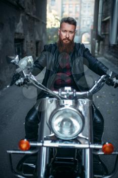 Bearded biker in leather jacket sitting on classical chopper, front view. Vintage bike, rider on motorcycle