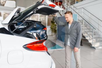 Man looks at the trunk of new car in showroom. Male customer choosing vehicle in dealership, automobile sale, auto purchase