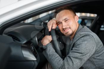 Happy man sitting in new car, showroom. Male customer choosing vehicle in dealership, automobile sale, auto purchase
