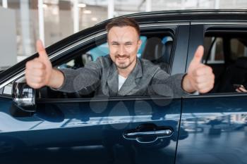 Man sitting in new car and shows thumbs up, showroom. Customer choosing vehicle in dealership, automobile sale, auto purchase