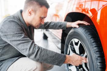 Man looks on wheels of new car in showroom. Male customer buying vehicle in dealership, automobile sale, auto purchase