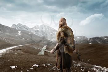 Viking with axes in hands dressed in traditional nordic clothes standing at the rocky mountains. Scandinavian ancient warrior