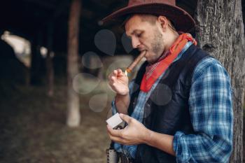 Brutal cowboy lights a cigar with matches, texas ranch on background, western. Vintage male person relax on farm, wild west lifestyle