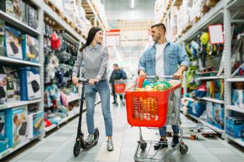 Couple in shop, husband with cart full of goods and wife on the scooter.  Man and woman in a supermarket, family shopping