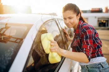 Young woman with sponge scrubbing vehicle glass with foam, car wash. Lady on self-service automobile washing. Outdoor carwash