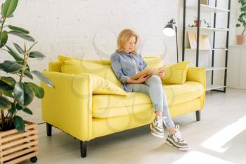 Young woman reading a book on cozy yellow couch, living room in white tones on background. Attractive female person with magazine sitting on sofa at home
