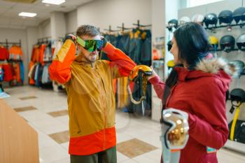 Couple at the showcase trying on mask for ski or snowboarding, shopping in sports shop. Winter season extreme lifestyle, active leisure store, customers buying glasses, protect equipment