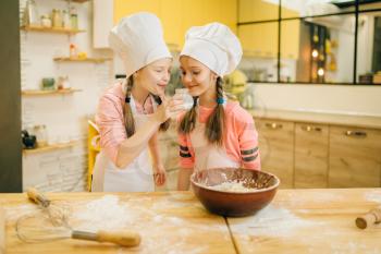 Two little girls cooks in caps sniffing sweet vanilla powder, cookies preparation on the kitchen. Kids cooking pastry, children chefs preparing cake