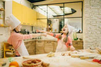 Two little girls chefs are laughing at each other, cookies preparation on the kitchen. Kids cooking pastry and having fun, children preparing cake